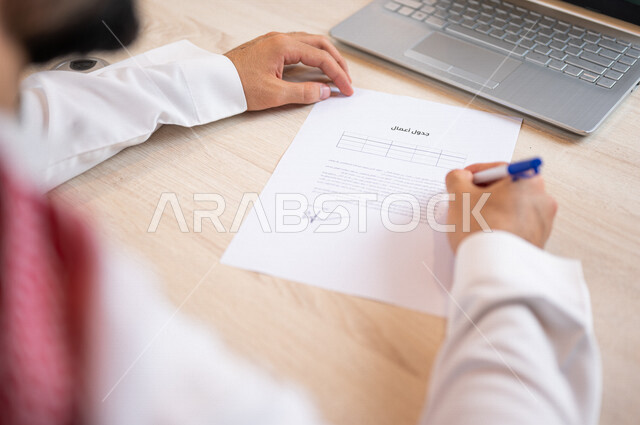 رجل عربي خليجي سعودي يجلس أمام الحاسوب المحمول ويقوم بتجهيز جدول الأعمال، سكرتير يدير الاعمال الورقية، تنظيم جدول الاعمال والمهام، أعمال مكتبية