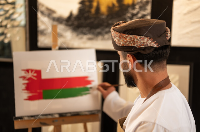 إحياء ذكرى تأسيس الدولة 1965، ممارسة الهوايات المفضلة والفنون اليدوية، مفهوم الموهبة و الإبداع وحب الوطن والانتماء إليه، الاحتفال باليوم الوطني العماني 18 نوفمبرفي ورشة عمل فنية، رسام عربي خليجي عماني يرتدي الدشداشة والعمامة مع مريلة الرسم يقوم برسم علم سلطنة عمان