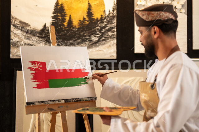 الاحتفال باليوم الوطني العماني 18 نوفمبرفي ورشة عمل فنية، رسام عربي خليجي عماني يرتدي الدشداشة والعمامة مع مريلة الرسم يقوم برسم علم سلطنة عمان، إحياء ذكرى تأسيس الدولة 1965، ممارسة الهوايات المفضلة والفنون اليدوية، مفهوم الموهبة و الإبداع وحب الوطن والانتماء إليه