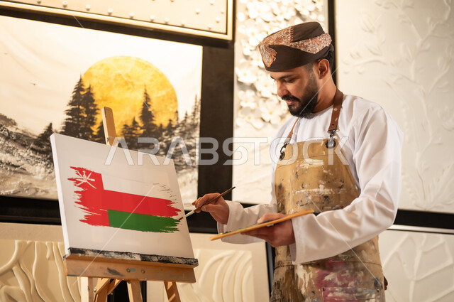 الاحتفال باليوم الوطني العماني 18 نوفمبرفي ورشة عمل فنية، رسام عربي خليجي عماني يرتدي الدشداشة والعمامة مع مريلة الرسم يقوم برسم علم سلطنة عمان، إحياء ذكرى تأسيس الدولة 1965، ممارسة الهوايات المفضلة والفنون اليدوية، مفهوم الموهبة و الإبداع وحب الوطن والانتماء إليه