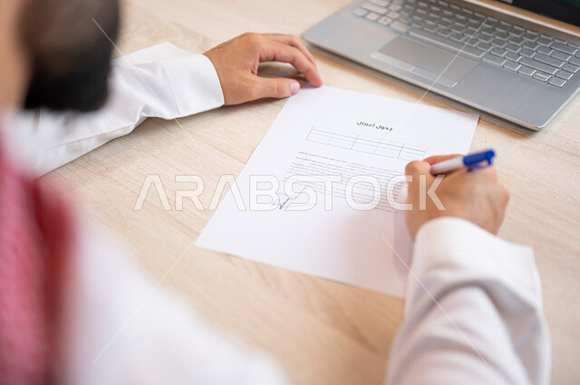 A Saudi Arabian Gulf man sitting in front of a laptop and preparing the agenda, a secretary who manages paperwork, organizing the agenda and tasks, office work