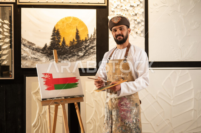 الاحتفال باليوم الوطني العماني 18 نوفمبرفي ورشة عمل فنية، رسام عربي خليجي عماني يرتدي الدشداشة والعمامة مع مريلة الرسم يقوم برسم علم سلطنة عمان، إحياء ذكرى تأسيس الدولة 1965، ممارسة الهوايات المفضلة والفنون اليدوية، مفهوم الموهبة و الإبداع وحب الوطن والانتماء إليه