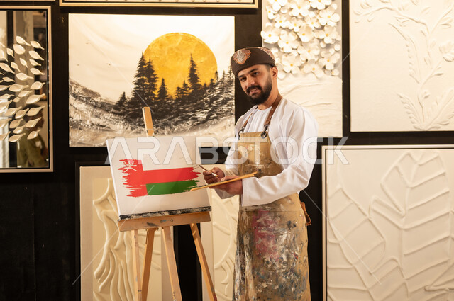اليوم الوطني العماني 18 نوفمبر، ورشة عمل فنية، رسام عربي خليجي عماني يرتدي الدشداشة والعمامة مع مريلة الرسم يقوم برسم علم سلطنة عمان، إحياء ذكرى تأسيس الدولة 1965، ممارسة الهوايات المفضلة والفنون اليدوية، مفهوم الموهبة و الإبداع وحب الوطن والانتماء إليه
