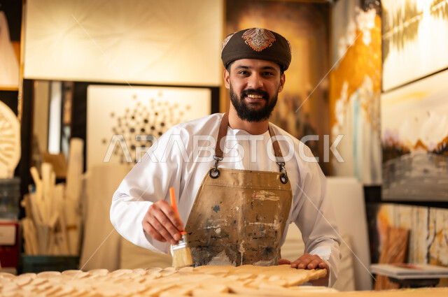 منحوتات وزخارف وأعمال فنية يدوية، حرف ومهن شعبية تراثية، معرض للمواهب والفنون التشكيلية الصناعية، رجل نحات عربي خليجي عماني يرتدي الدشداشة والعمامة التقليدية مع المريلة  يقوم بالنحت والرسم على الخشب في ورشة العمل