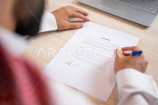 رجل عربي خليجي سعودي يجلس أمام الحاسوب المحمول ويمسك بيده قلم، القيام بقراءة العرض الفني، سكرتير يدير الاعمال الورقية، التحقق من بنود العرض، مفهوم التوظيف والعمل، أعمال مكتبية