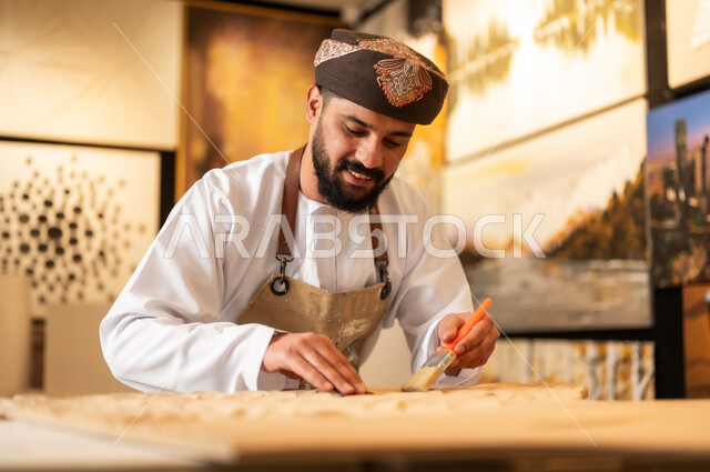 زخارف ومنحوتات وأعمال فنية يدوية، حرف ومهن شعبية تراثية، معرض للمواهب والفنون التشكيلية الصناعية، رجل نحات عربي خليجي عماني يرتدي الدشداشة والعمامة التقليدية مع المريلة  يقوم بالنحت والرسم على الخشب في ورشة العمل