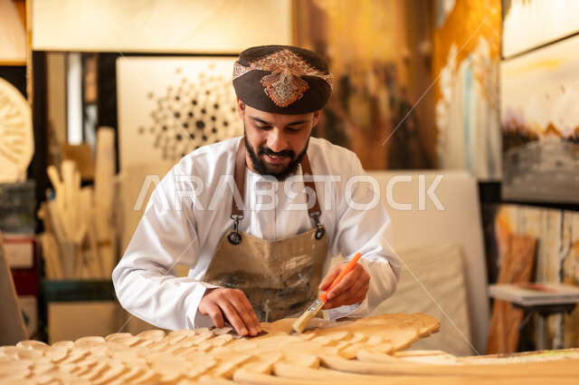 منحوتات وزخارف وأعمال فنية يدوية، حرف ومهن شعبية تراثية، معرض للمواهب والفنون التشكيلية الصناعية، رجل نحات عربي خليجي عماني يرتدي الدشداشة والعمامة التقليدية مع المريلة  يقوم بالنحت والرسم على الخشب في ورشة العمل