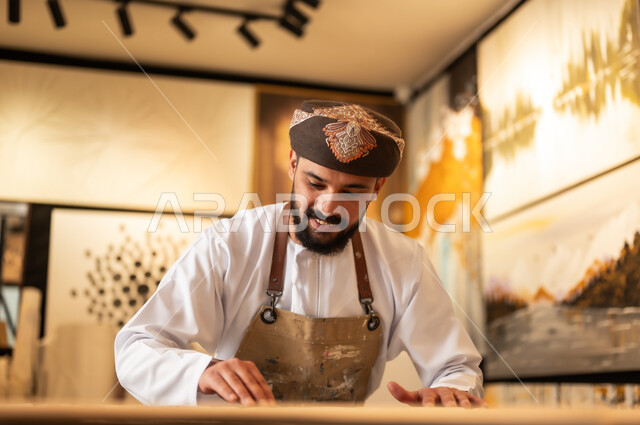 زخارف ومنحوتات وأعمال فنية يدوية، حرف ومهن شعبية تراثية، معرض للمواهب والفنون التشكيلية الصناعية، رجل نحات عربي خليجي عماني يرتدي الدشداشة والعمامة التقليدية مع المريلة  يقوم بالنحت والرسم على الخشب في ورشة العمل