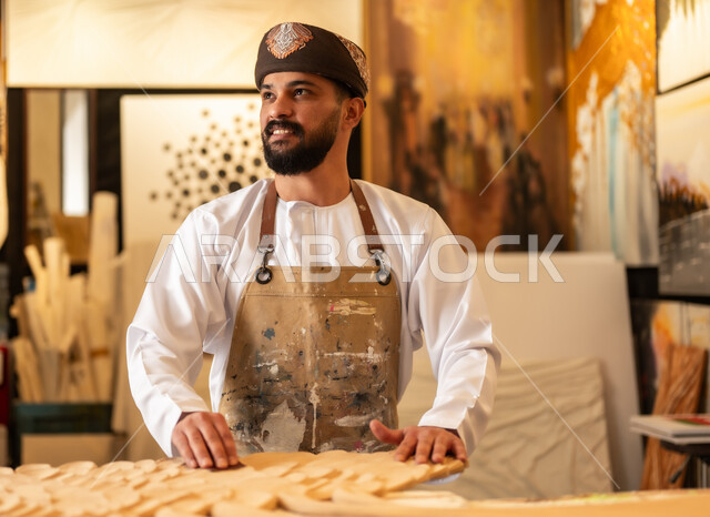زخارف ومنحوتات وأعمال فنية يدوية، حرف ومهن شعبية تراثية، معرض للمواهب والفنون التشكيلية الصناعية، رجل نحات عربي خليجي عماني يرتدي الدشداشة والعمامة التقليدية مع المريلة  يقوم بالنحت والرسم على الخشب في ورشة العمل