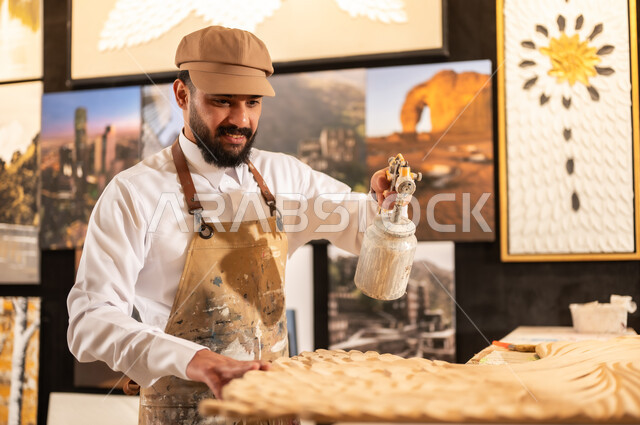 Traditional folk crafts and professions, an exhibition of talents, fine arts and handicrafts, a Saudi Arabian Gulf Arab sculptor wearing traditional thobe and apron spraying sculptures and coloring wood in the workshop, sculptures, decorations and handmade artworks