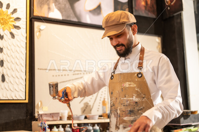 Traditional folk crafts and professions, an exhibition of talents, fine arts and handicrafts, a Saudi Arabian Gulf Arab sculptor wearing traditional thobe and apron spraying sculptures and coloring wood in the workshop, sculptures, decorations and handmade artworks