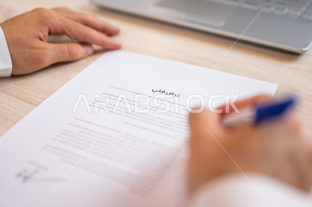 رجل عربي خليجي سعودي يجلس أمام الحاسوب المحمول ويمسك بيده قلم، القيام بقراءة العرض الفني، سكرتير يدير الاعمال الورقية، التحقق من بنود العرض، مفهوم التوظيف والعمل، أعمال مكتبية