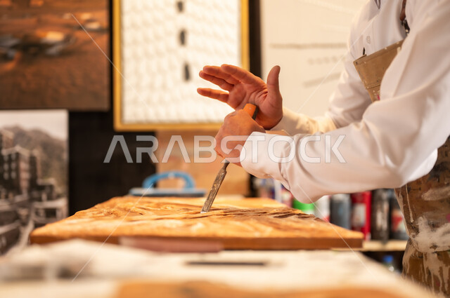 زخارف ومنحوتات وأعمال فنية يدوية، حرف ومهن شعبية تراثية، معرض للمواهب والفنون التشكيلية والصناعات اليدوية، رجل نحات عربي خليجي سعودي يرتدي الثوب التقليدية والمريلة  يقوم بالنحت والرسم على الخشب في ورشة العمل