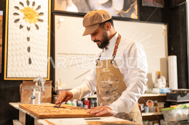 منحوتات وزخارف وأعمال فنية يدوية، حرف ومهن شعبية تراثية، معرض للمواهب والفنون التشكيلية والصناعات اليدوية، رجل نحات عربي خليجي سعودي يرتدي الثوب التقليدية والمريلة يقوم بالنحت والرسم على الخشب في ورشة العمل