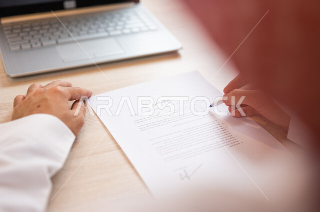 رجل عربي خليجي سعودي يجلس أمام الحاسوب المحمول ويمسك بيده قلم، القيام بقراءة العرض الفني، سكرتير يدير الاعمال الورقية، التحقق من بنود العرض، مفهوم التوظيف والعمل، أعمال مكتبية