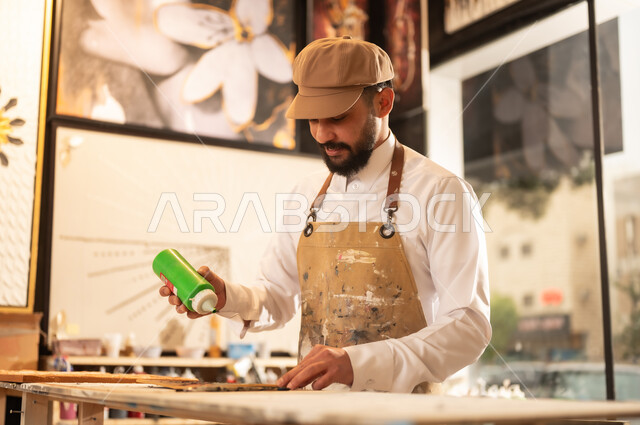 خلط الألوان الاساسية، مفهوم الموهبة و الإبداع, ورشة عمل فنية, رسام عربي خليجي سعودي يرتدي الثوب التقليدي ومريلة الرسم يقوم باستعمال اللون الاخضر, الشغف في ممارسة الهوايات المفضلة، استخدام أدوات و معدات الرسم و التلوين، تعابير وايماءات الاندماج والسرور