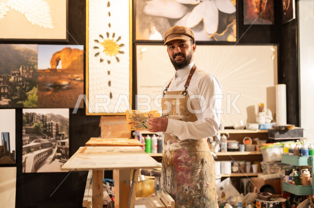 Using a brush to draw on a canvas, the concept of talent and creativity, an art workshop, a Saudi Arabian Gulf painter wearing traditional clothing and a painting apron sitting on a chair thinking about drawing something, passion for practicing favorite hobbies and handicrafts, expressions and gestures of integration and pleasure