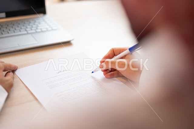 رجل عربي خليجي سعودي يجلس أمام الحاسوب المحمول ويمسك بيده قلم، القيام بقراءة العرض الفني، سكرتير يدير الاعمال الورقية، التحقق من بنود العرض، مفهوم التوظيف والعمل، أعمال مكتبية