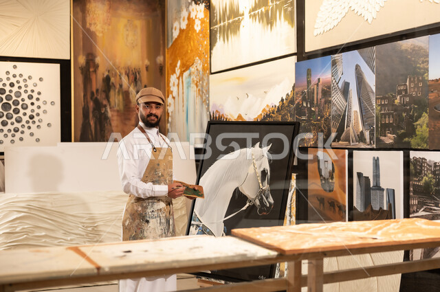تعابير وايماءات الاندماج والسرور، استخدام الريشة للرسم على اللوحة، مفهوم الموهبة و الإبداع، ورشة عمل فنية، رسام عربي خليجي سعودي يرتدي الثوب التقليدي ومريلة الرسم يستعد لرسم شيء ما، الشغف في ممارسة الهوايات المفضلة والفنون اليدوية