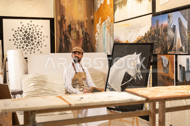 Using a brush to draw on a canvas, the concept of talent and creativity, an art workshop, a Saudi Arabian Gulf painter wearing traditional clothing and a painting apron sitting on a chair thinking about drawing something, passion for practicing favorite hobbies and handicrafts, expressions and gestures of integration and pleasure