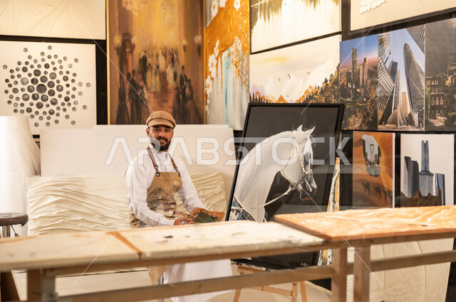 Using a brush to draw on a canvas, the concept of talent and creativity, an art workshop, a Saudi Arabian Gulf painter wearing traditional clothing and a painting apron sitting on a chair thinking about drawing something, passion for practicing favorite hobbies and handicrafts, expressions and gestures of integration and pleasure