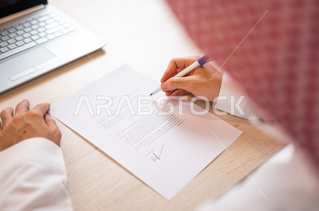 رجل عربي خليجي سعودي يجلس أمام الحاسوب المحمول ويمسك بيده قلم، القيام بقراءة العرض الفني، سكرتير يدير الاعمال الورقية، التحقق من بنود العرض، مفهوم التوظيف والعمل، أعمال مكتبية