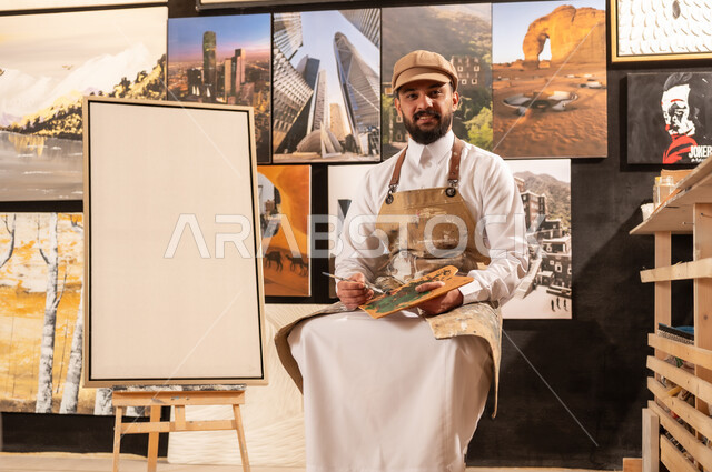 استخدام الريشة للرسم على اللوحة، مفهوم الموهبة و الإبداع, ورشة عمل فنية, رسام عربي خليجي سعودي يرتدي الثوب التقليدي ومريلة الرسم يجلس على الكرسي يفكر في رسم شيء ما, الشغف في ممارسة الهوايات المفضلة والفنون اليدوية، تعابير وايماءات الاندماج والسرور