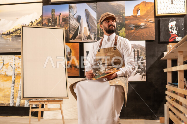 استخدام الريشة للرسم على اللوحة، مفهوم الموهبة و الإبداع, ورشة عمل فنية, رسام عربي خليجي سعودي يرتدي الثوب التقليدي ومريلة الرسم يجلس على الكرسي يفكر في رسم شيء ما, الشغف في ممارسة الهوايات المفضلة والفنون اليدوية، تعابير وايماءات الاندماج والسرور