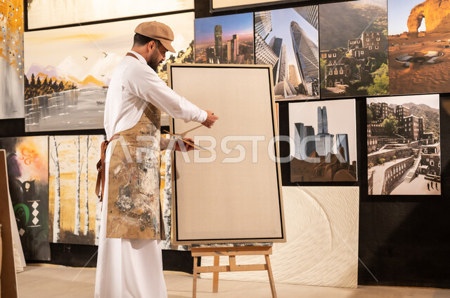 تعابير وايماءات الاندماج والسرور، استخدام الريشة للرسم على اللوحة، مفهوم الموهبة و الإبداع، ورشة عمل فنية، رسام عربي خليجي سعودي يرتدي الثوب التقليدي ومريلة الرسم يستعد لرسم شيء ما، الشغف في ممارسة الهوايات المفضلة والفنون اليدوية