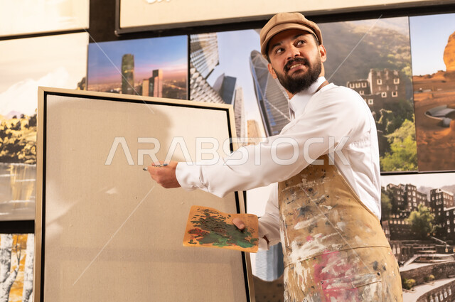 Expressions and gestures of integration and pleasure, using a brush to draw on a canvas, the concept of talent and creativity, an art workshop, a Saudi Arabian Gulf artist wearing traditional clothing and a painting apron preparing to draw something, passion for practicing favorite hobbies and handicrafts