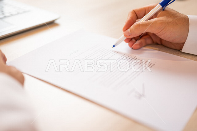 رجل عربي خليجي سعودي يجلس أمام الحاسوب المحمول ويمسك بيده قلم، القيام بقراءة العرض الفني، سكرتير يدير الاعمال الورقية، التحقق من بنود العرض، مفهوم التوظيف والعمل، أعمال مكتبية