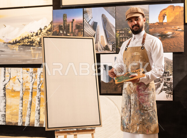 تعابير وايماءات الاندماج والسرور، استخدام الريشة للرسم على اللوحة، مفهوم الموهبة و الإبداع، ورشة عمل فنية، رسام عربي خليجي سعودي يرتدي الثوب التقليدي ومريلة الرسم يستعد لرسم شيء ما، الشغف في ممارسة الهوايات المفضلة والفنون اليدوية