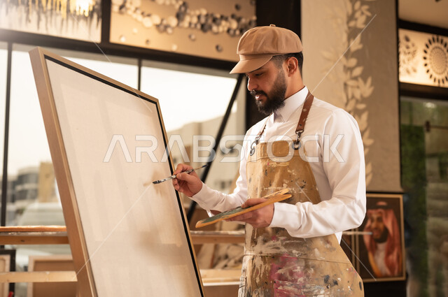 تعابير وايماءات الاندماج والسرور، استخدام الريشة للرسم على اللوحة، مفهوم الموهبة و الإبداع، ورشة عمل فنية، رسام عربي خليجي سعودي يرتدي الثوب التقليدي ومريلة الرسم يستعد لرسم شيء ما، الشغف في ممارسة الهوايات المفضلة والفنون اليدوية