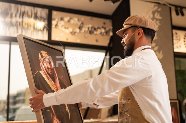 الإبداع والموهبة في الفنون اليدوية، تعابير السرور والسعادة، مفهوم الحب والانتماء للوطن, ورشة عمل فنية سعودية, رسام عربي خليجي سعودي يرتدي الثوب التقليدي ومريلة الرسم يقدم فنون الرسم و التلوين لصورة الملك عبد العزيز آل سعود