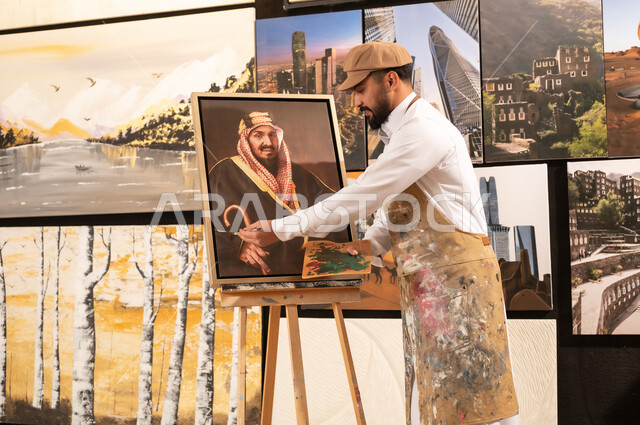 الموهبة و الإبداع في الفنون اليدوية، تعابير السرور والسعادة، مفهوم الحب والانتماء للوطن, ورشة عمل فنية سعودية, رسام عربي خليجي سعودي يرتدي الثوب التقليدي ومريلة الرسم يقدم فنون الرسم و التلوين لصورة الملك عبد العزيز آل سعود