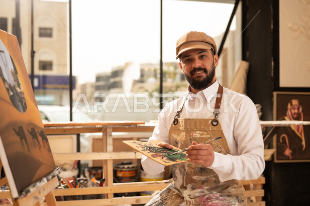 Practicing the hobby of handicrafts, passion for drawing animals, a Saudi Arabian Gulf artist wearing traditional dress and a painting apron drawing camels in the desert and landmarks of Riyadh, an exhibition of drawings and paintings in the Saudi capital, the concept of talent and creativity