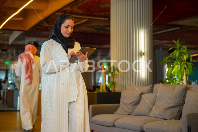  ادارة مهام العمل اليومية، مهن ووظائف مكتبية، بيئة العمل المريحة في الشركات السعودية, سيدة اعمال عربية سعودية خليجية ترتدي الحجاب والعباءة تحمل بيدها اوراق تتجول داخل مقر الشركة