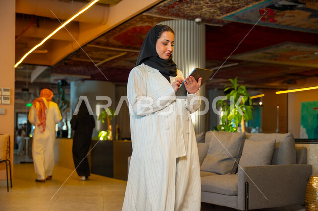  ادارة مهام العمل اليومية، مهن ووظائف مكتبية، بيئة العمل المريحة في الشركات السعودية, سيدة اعمال عربية سعودية خليجية ترتدي الحجاب والعباءة تحمل بيدها اوراق تتجول داخل مقر الشركة