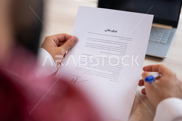رجل عربي خليجي سعودي يجلس أمام الحاسوب المحمول، القيام بقراءة بنود العرض، التحقق والتدقيق في العرض المالي، الموافقة على العرض المالي