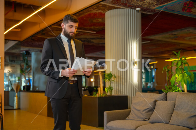  ادارة مهام العمل اليومية،  مهن ووظائف مكتبية، بيئة العمل المريحة في الشركات السعودية, رجل اعمال عربي سعودي خليجي يرتدي البدلة الرسمية يحمل بيده اوراق يتجول داخل مقر الشركة
