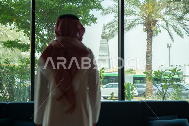 الفن الهندسي المعماري للأبراج و ناطحات السحاب في العاصمة، صورة من الخلف لرجل عربي خليجي سعودي يرتدي الشماغ والثوب التقليدي يقوم بمشاهدة برج الفيصلية في مدينة الرياض، الواجهات الزجاجية الكبيرة في شركات المملكة العربية السعودية، معلم سياحي مشهور