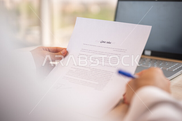 رجل عربي خليجي سعودي يجلس امام الحاسوب المحمول وبيده أوراق خاصة بعقد عمل، قراءة البنود الخاصة بالعقد، التحقق والتدقيق في شروط عقد الايجار، الموافقة على الشروط، التوقيع على العقود