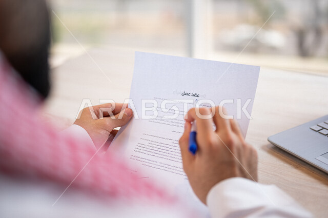 رجل عربي خليجي سعودي يجلس امام الحاسوب المحمول وبيده أوراق خاصة بعقد عمل، قراءة البنود الخاصة بالعقد، التحقق والتدقيق في شروط عقد الايجار، الموافقة على الشروط، التوقيع على العقود