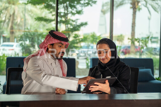 Business development and problem solving, Saudi office jobs and professions, exchanging opinions and experiences, a meeting of a Saudi Gulf Arab businessman and woman at the company headquarters, managing work using a tablet, discussing and distributing work tasks, the Saudi work environment, unity and teamwork, achieving company goals.