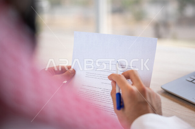 رجل عربي خليجي سعودي يجلس امام الحاسوب المحمول وبيده أوراق خاصة بعقد عمل، قراءة البنود الخاصة بالعقد، التحقق والتدقيق في شروط عقد الايجار، الموافقة على الشروط، التوقيع على العقود