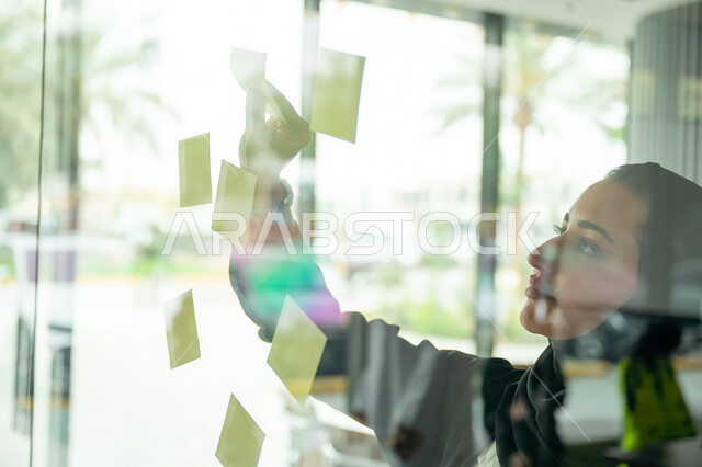 كتابة وتدوين شيء ما, أوراق الملاحظات الملونة المعلقة, بيئة العمل في الشركات السعودية, مهن وأعمال مكتبية, صورة مقربة لامرأة عربية خليجية سعودية ترتدي الحجاب والعباءة تقوم بلصق ورقة ملاحظة على واجهة زجاجية داخل المكتب