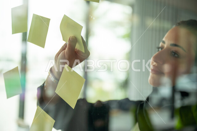 كتابة وتدوين شيء ما, أوراق الملاحظات الملونة المعلقة, بيئة العمل في الشركات السعودية, مهن وأعمال مكتبية, صورة مقربة لامرأة عربية خليجية سعودية ترتدي الحجاب والعباءة تقوم بلصق ورقة ملاحظة على واجهة زجاجية داخل المكتب