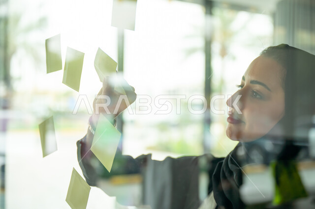 كتابة وتدوين شيء ما, أوراق الملاحظات الملونة المعلقة, بيئة العمل في الشركات السعودية, مهن وأعمال مكتبية, صورة مقربة لامرأة عربية خليجية سعودية ترتدي الحجاب والعباءة تقوم بلصق ورقة ملاحظة على واجهة زجاجية داخل المكتب
