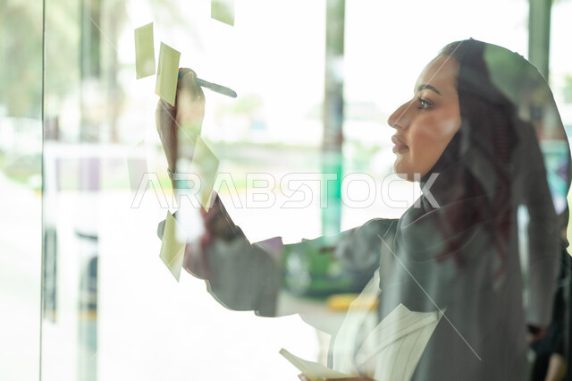 كتابة وتدوين شيء ما, أوراق الملاحظات الملونة المعلقة, بيئة العمل في الشركات السعودية, مهن وأعمال مكتبية, صورة مقربة لامرأة عربية خليجية سعودية ترتدي الحجاب والعباءة تقوم بلصق ورقة ملاحظة على واجهة زجاجية داخل المكتب