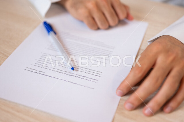 رجل عربي خليجي سعودي امامه أوراق خاصة بعقد وفوقها قلم، سكرتير يدير الاعمال الورقية، قراءة البنود الخاصة بالعقد، التحقق والتدقيق في شروط العقد، الموافقة على الشروط